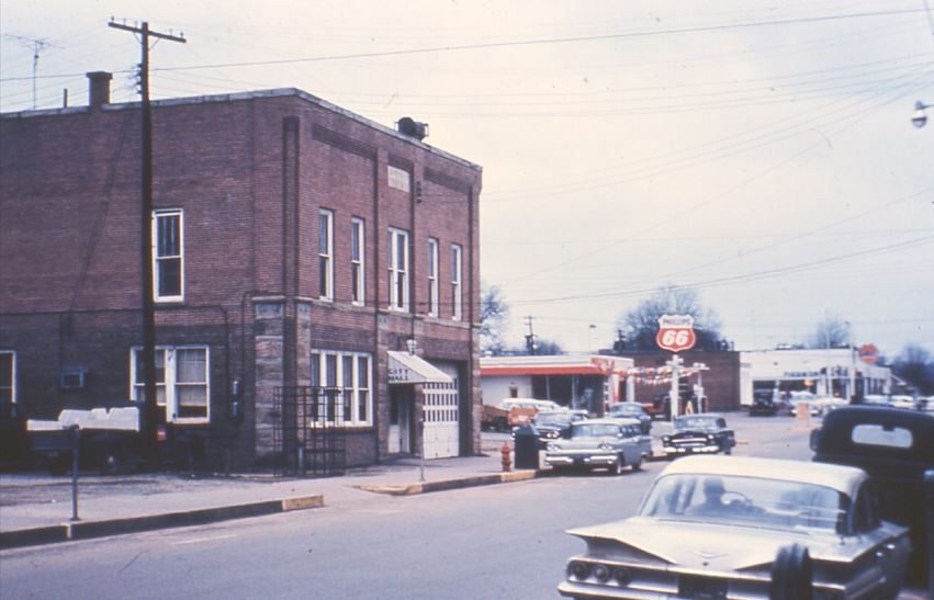 The two-story brick City Hall with a fire bay door and a Phillips 66 station beyond