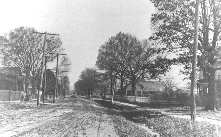 A dirt East Ladiga Street lined with oaks, picket fences, and utility poles